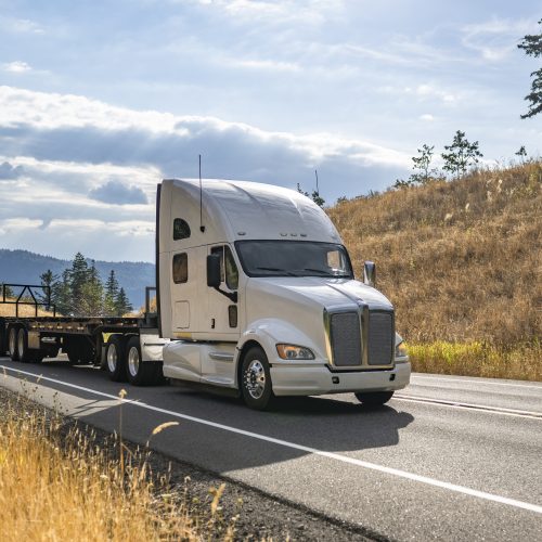 Big rig white semi truck transporting two empty flat bed semi trailer running on the winding road to warehouse for the next load